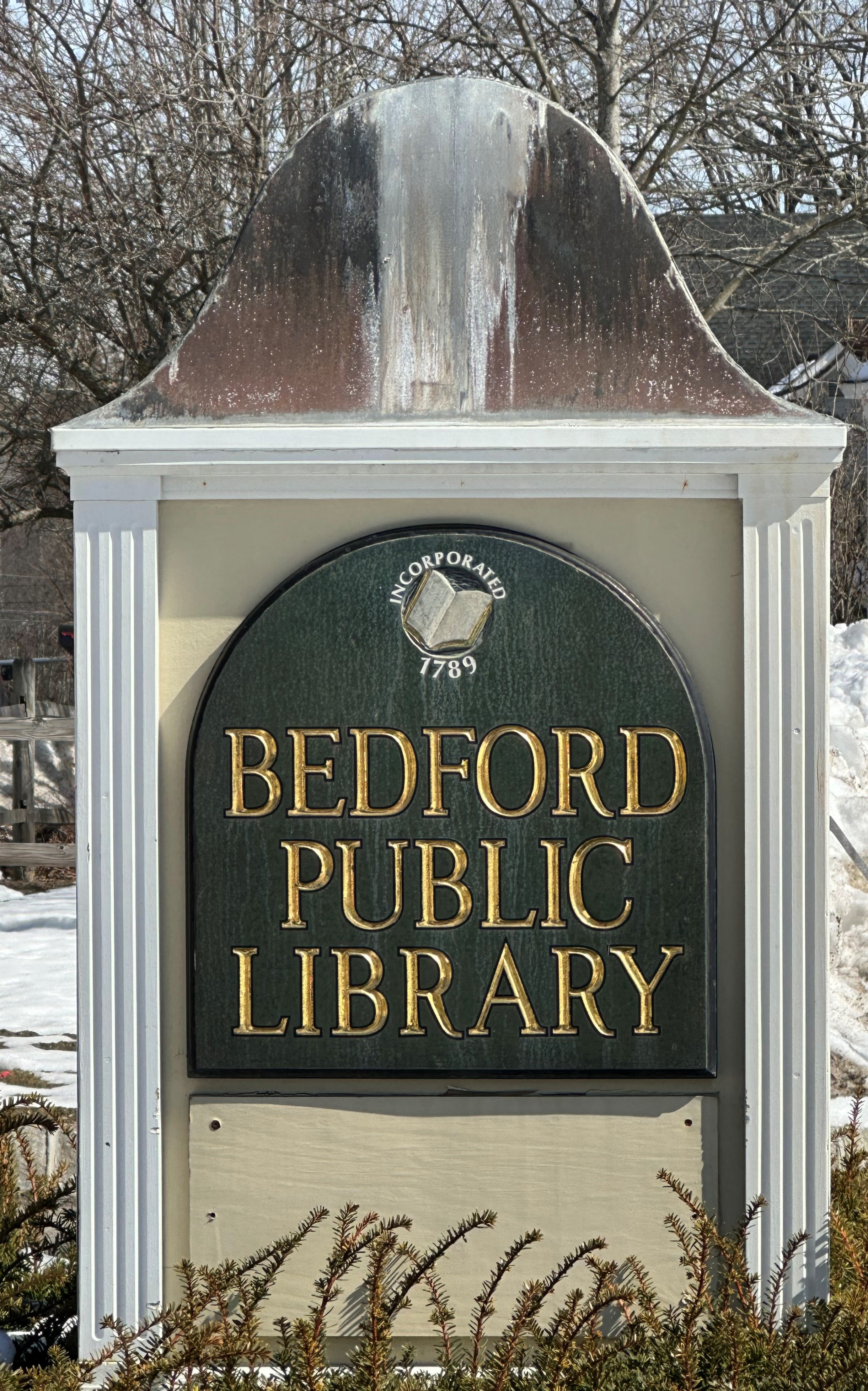 Bedford Public Library town sign, gold lettering on a greenish-black arch shaped sign seated within a square sign with white borders and a snowy background, presented by New Hampshire personal injury lawyer Attorney Buckley