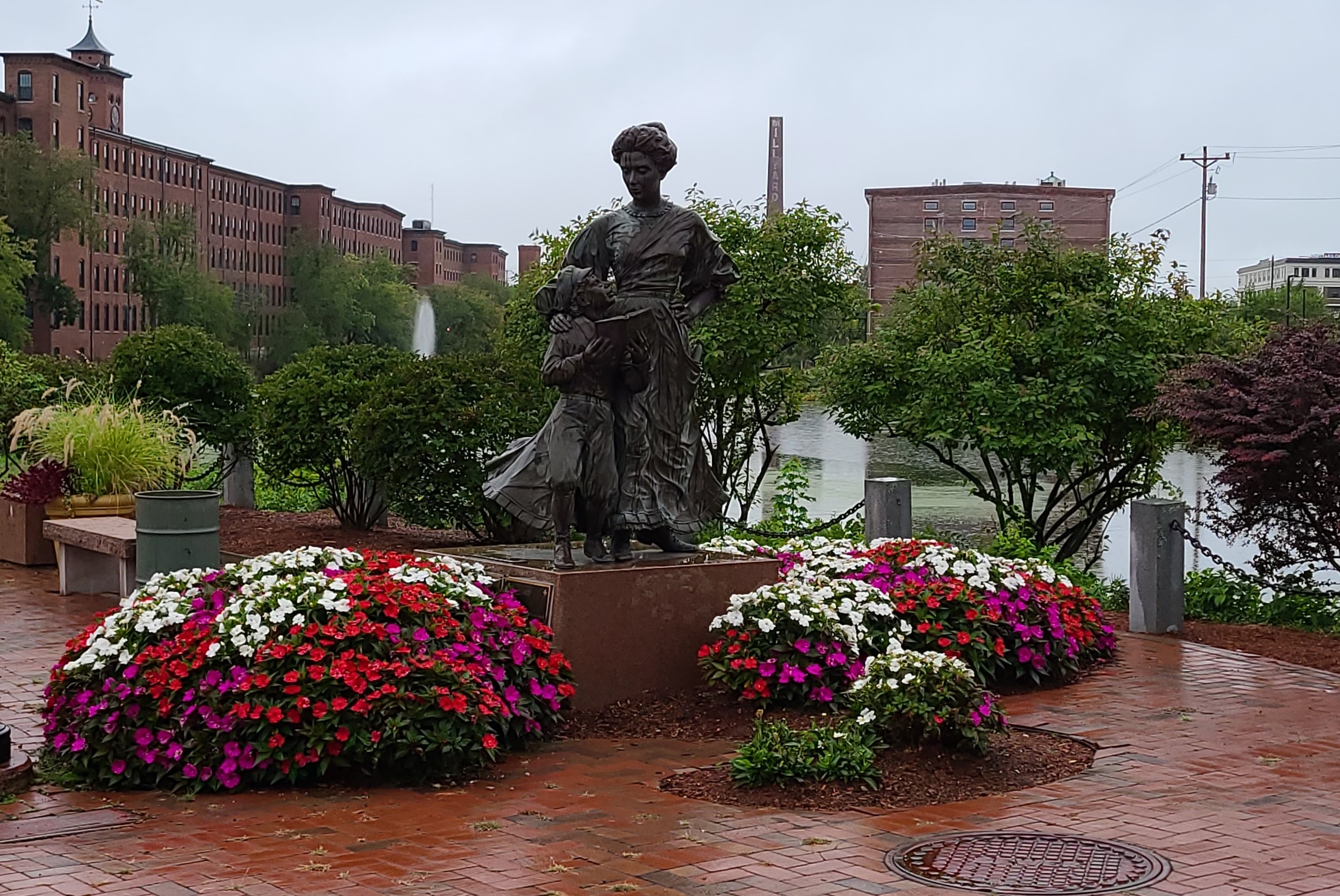 Photo of statue of a woman in dress and a boy looking up at her with an open book in the downtown Nashua garden, flowers blooming around the base of the statue are red, purple, and white, brick buildings populate the background of the photo, presented by Nashua personal injury lawyer Attorney Buckley