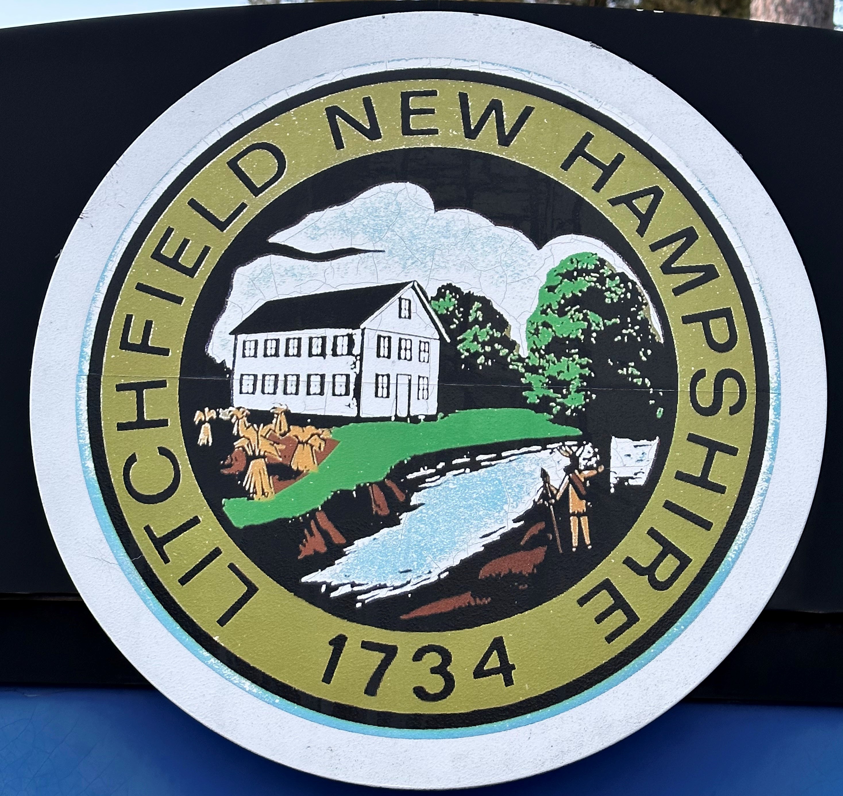 Litchfield town crest is a gold circle containing the words Litchfield New Hampshire 1734 , and inside the circle is an illustrated picture of a farm with a river and a man with a spear next to a tree, presented by New Hampshire personal injury law firm Buckley Law Offices.
