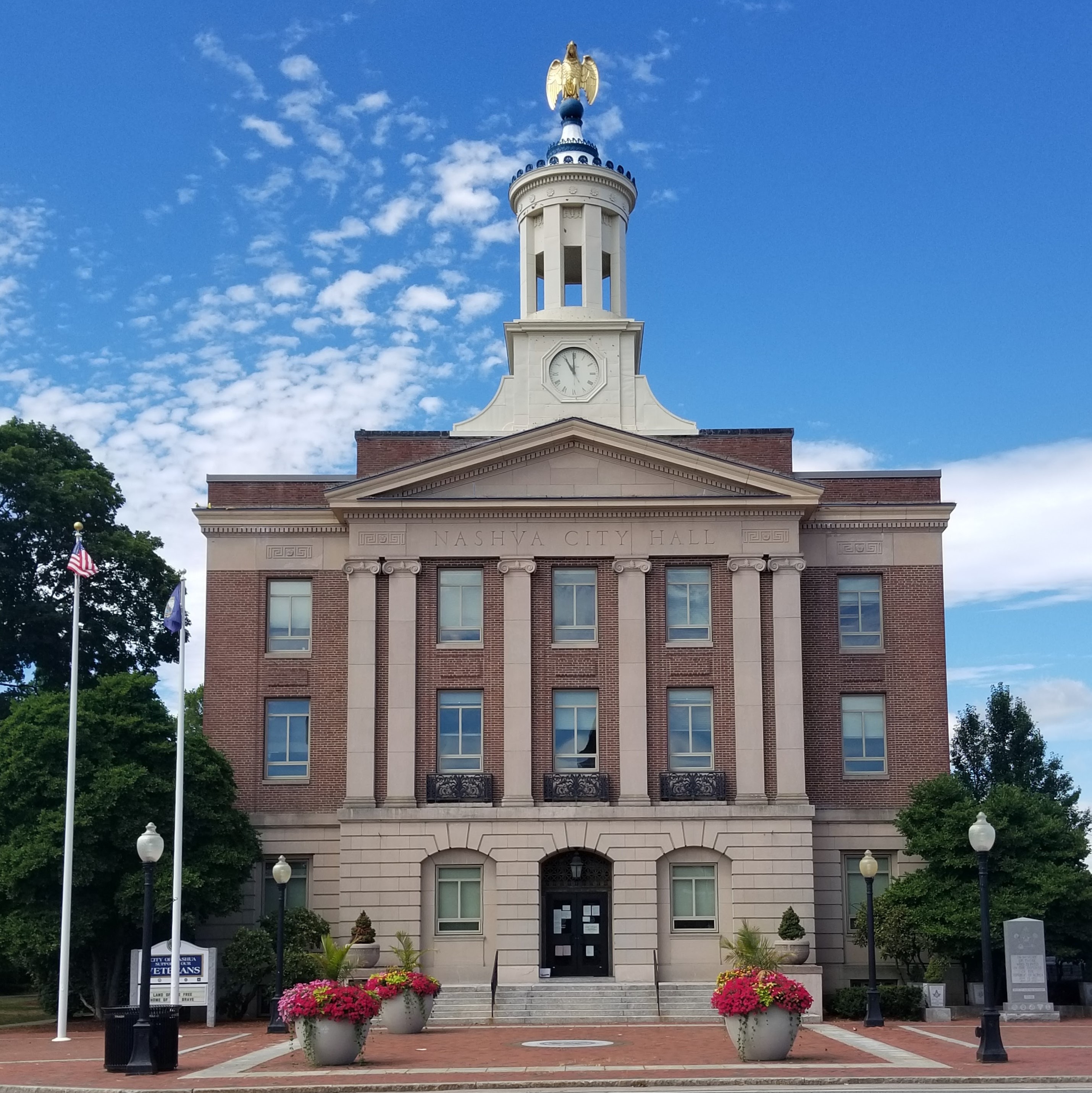 This is a photo of Nashua's city hall building. The photo is taken from the sidewalk, and the building is in the foreground of a mostly blue, slightly cloudy sky. The building is brick with beige cement bricks and columns accentuating the center of the building. At the top is a large clock, column accents, a dome, and a golden eagle. To the left is the American and New Hampshire state flags. New Hampshire personal injury lawyer Attorney Buckley features this photo.