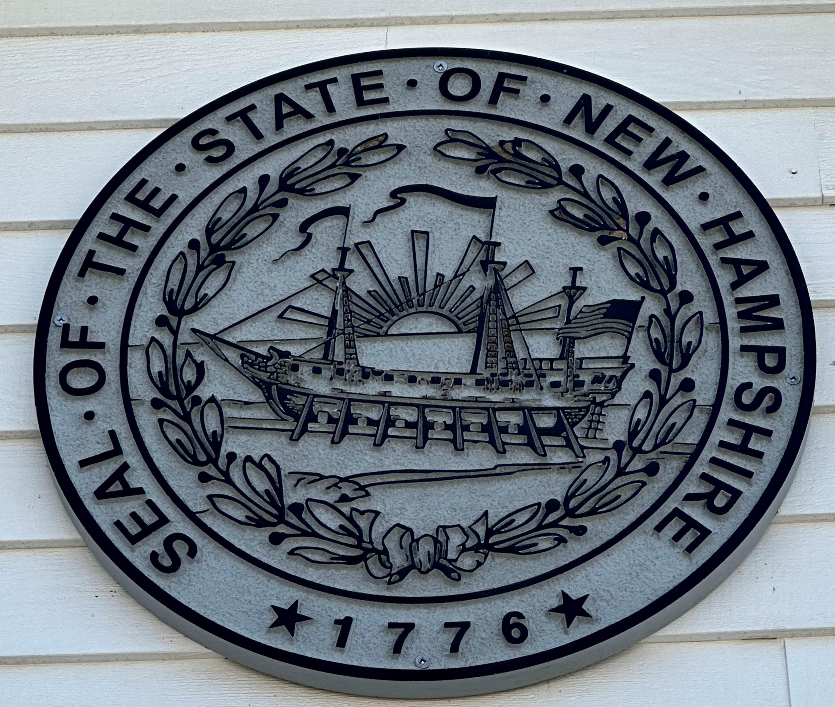 This is a photo of the seal of the state of New Hampshire, featured by personal injury lawyer Attorney Buckley. It is an oval grey plaque on a white wooden paneled wall. Words encircle a wreath that encircles an illustration of a sunrise behind a boat and an American flag. The words read: Seal of the State of New Hampshire, 1776, with a star on either side of the year.