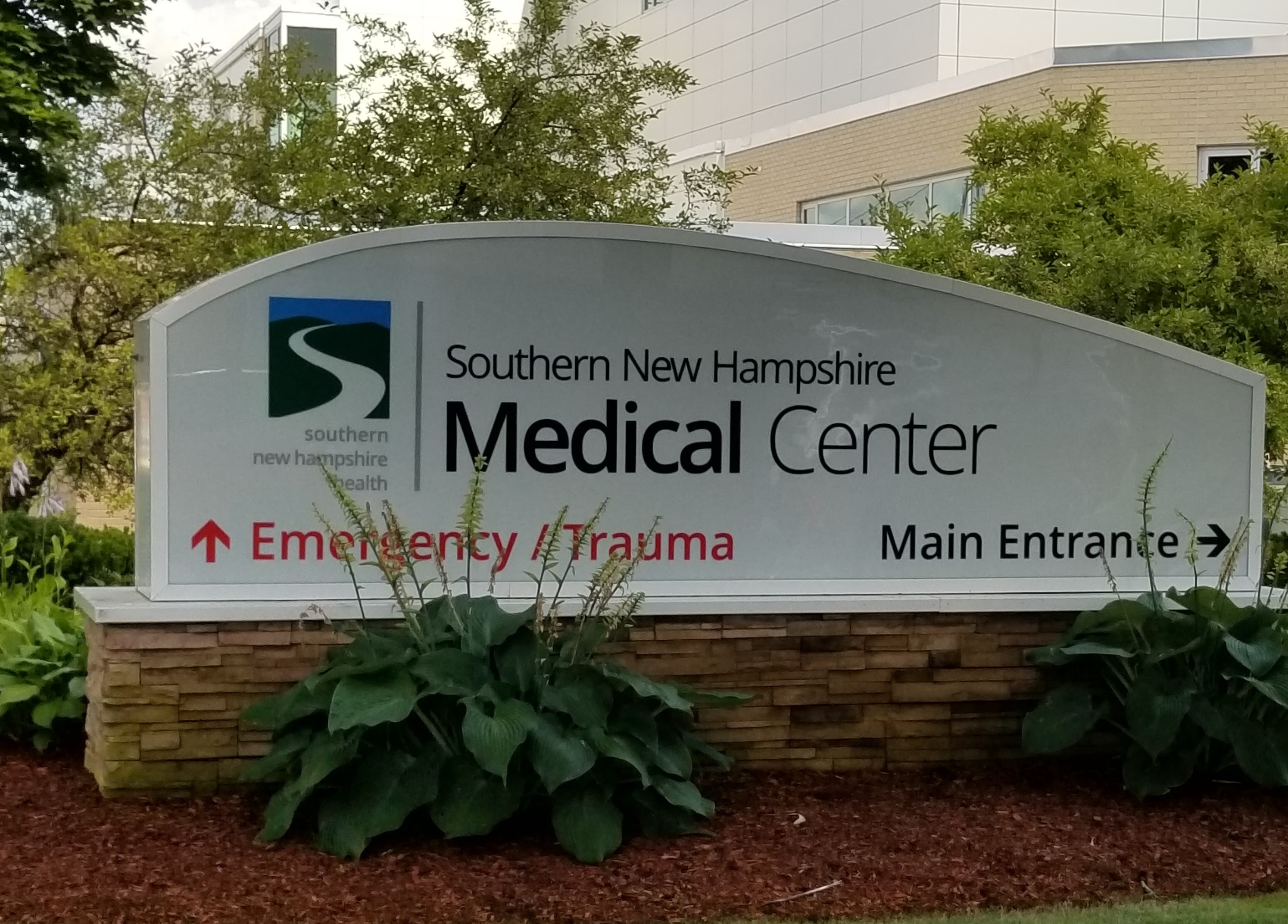 This is a photo of a sign at the Southern New Hampshire Medical Center. It directs drivers straight to Emergency/Trauma, highlighted with a red text on the sign's white background. On the right, it directs drivers to the right towards the main entrance in blue text. In blue text on the top right, the sign reads Southern New Hampshire Medical Center; a wavy logo with the same text appears on the left of the sign. The sign sits atop a beige brick base. Two low shrubs are in front of the sign, and behind the sign are some short trees and the medical center. New Hampshire personal injury lawyer Attorney Buckley features this photo.